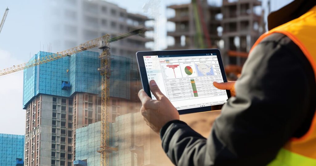 A person in a safety vest uses a tablet displaying data charts at a construction site with cranes and tall buildings in the background.