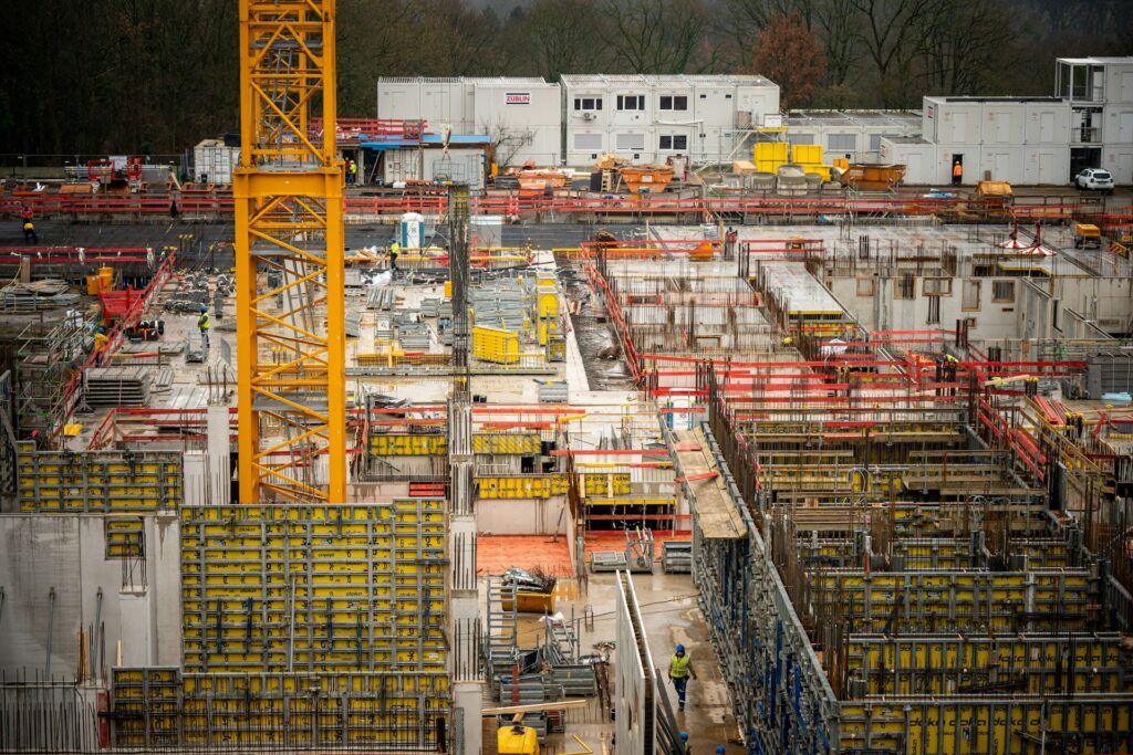A busy construction site with yellow cranes, concrete walls, scaffolding, and workers in safety vests. Portable buildings are in the background, surrounded by trees and construction materials.
