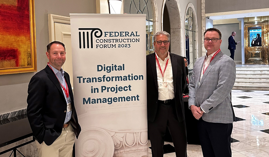 Three men in business attire stand beside a sign reading “Federal Construction Forum 2023: Digital Transformation in Project Management” in a marble-floored lobby.