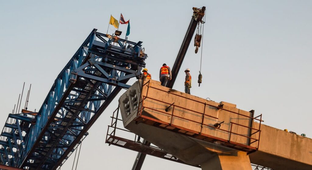 Construction workers in safety vests work on a bridge under construction, using cranes and heavy machinery, with flags on top of the structure against a clear sky.