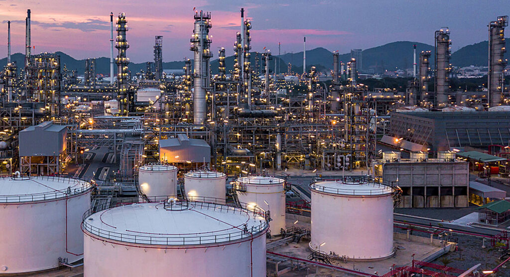 A large oil refinery at dusk with tall industrial towers, cylindrical storage tanks, and bright lights, set against a backdrop of hills and a purple sky.