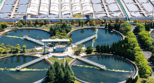 Aerial view of a modern water treatment plant with four circular clarifier tanks, surrounded by greenery and a large building with solar panels in the background.