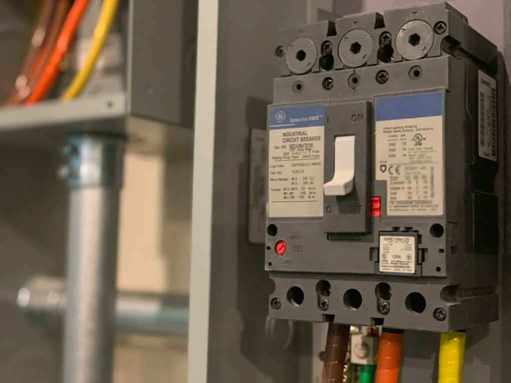 Close-up of an industrial circuit breaker with colored wires connected at the bottom, mounted on a gray electrical panel, and labels visible on the device.