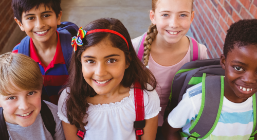 Five smiling children with backpacks stand close together outdoors by a brick wall, looking up at the camera. The group is diverse, and they appear to be ready for school.