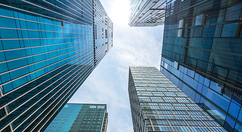 View looking up at modern glass skyscrapers from street level, with blue sky and sunlight visible between the tall buildings.