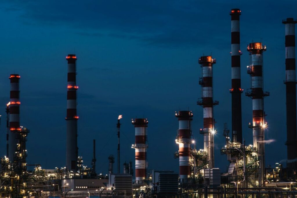 Industrial refinery at night with tall smokestacks and lights, emitting small amounts of smoke against a dark blue sky.