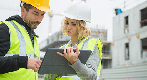 Two construction workers in safety vests and hard hats look at a tablet together at a building site with unfinished structures in the background.