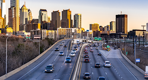 Cars travel on a busy multi-lane highway heading toward a city skyline with tall buildings in the background during sunset.