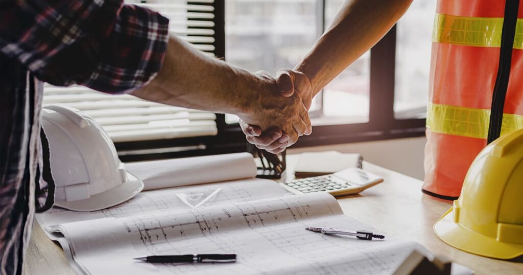 Two people shake hands over a desk with blueprints, pens, safety helmets, and a calculator, suggesting a construction or engineering agreement.