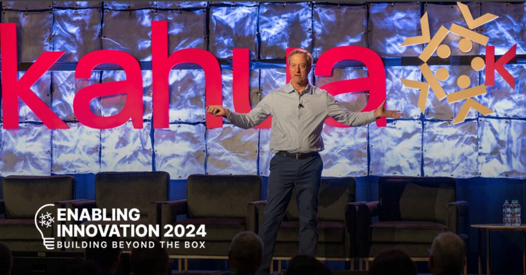 A man speaks onstage at Enabling Innovation 2024, with large red “kahua” text and logo behind him, and three empty chairs and bottled water to the side.