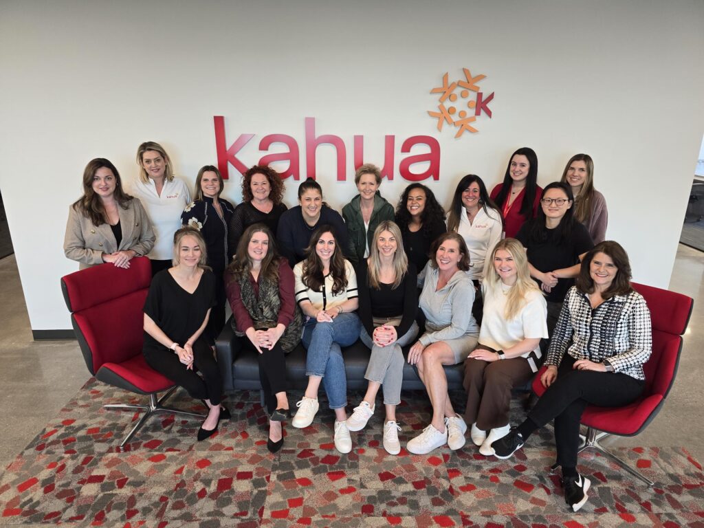 Twenty women pose together in front of a wall with the kahua logo, seated and standing in two rows on red chairs and a sofa, smiling at the camera in a modern office space.
