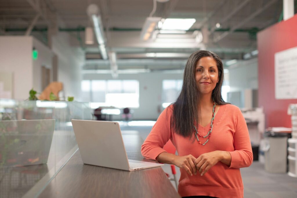 Woman with long dark hair wearing an orange sweater stands in a modern office by a laptop, smiling at the camera. Bright open workspace with plants and an industrial ceiling in the background.