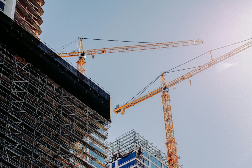 Two yellow construction cranes stand tall beside partially built skyscrapers covered in scaffolding, against a clear blue sky with sunlight shining from the top right corner.