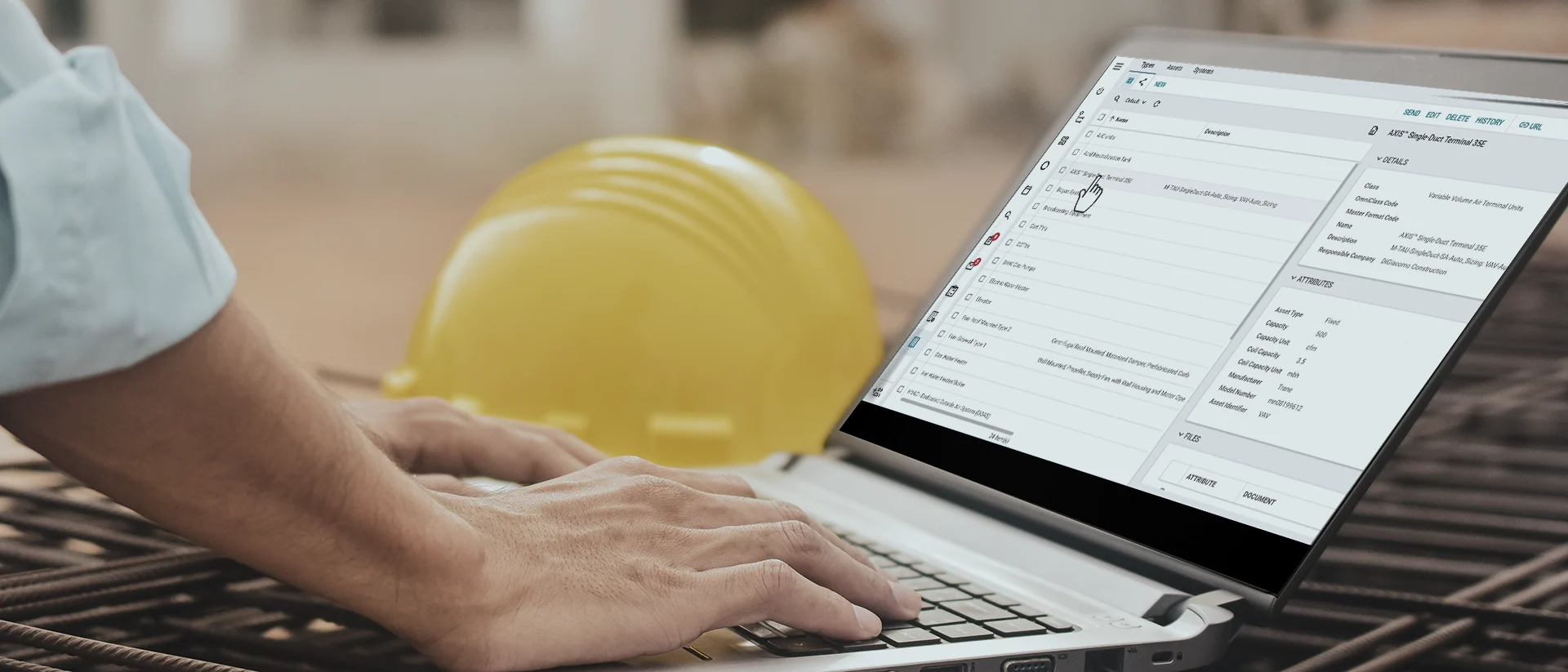 A person types on a laptop displaying a data entry form, with a yellow construction helmet in the background. The scene suggests work related to construction or project management.