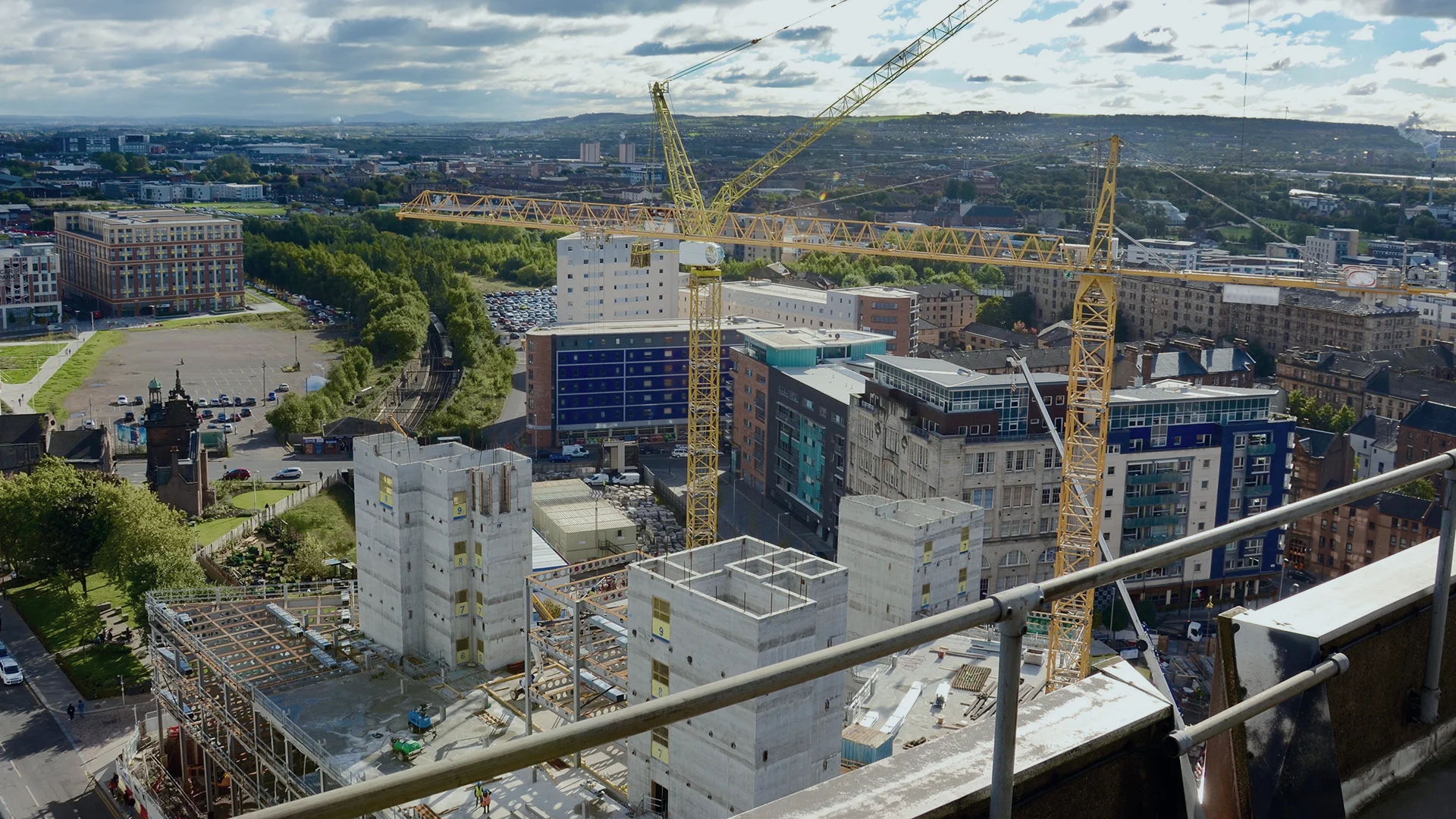 Aerial view of a city construction site with cranes and partially built concrete structures, surrounded by buildings, roads, and greenery under a cloudy sky.