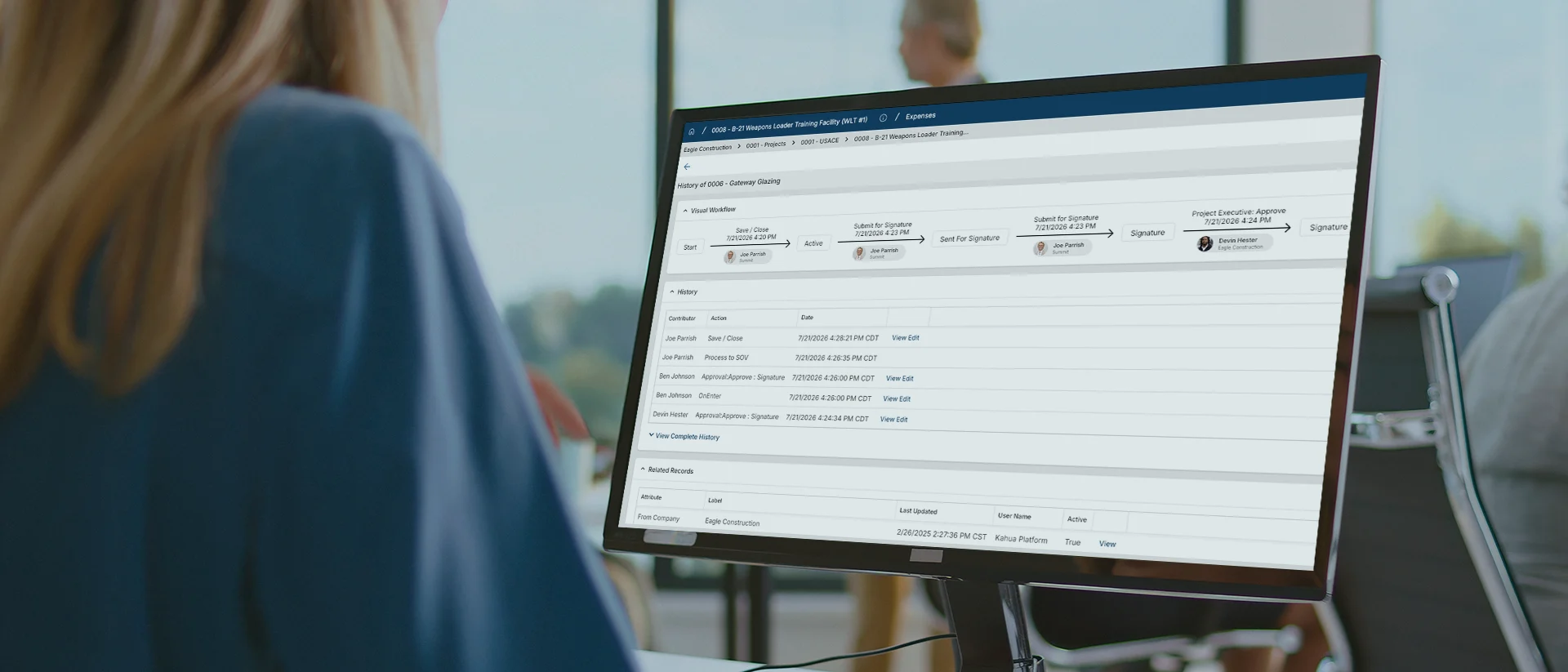A person sits at a desk looking at a computer monitor displaying a software interface with tables and search options in an office setting.