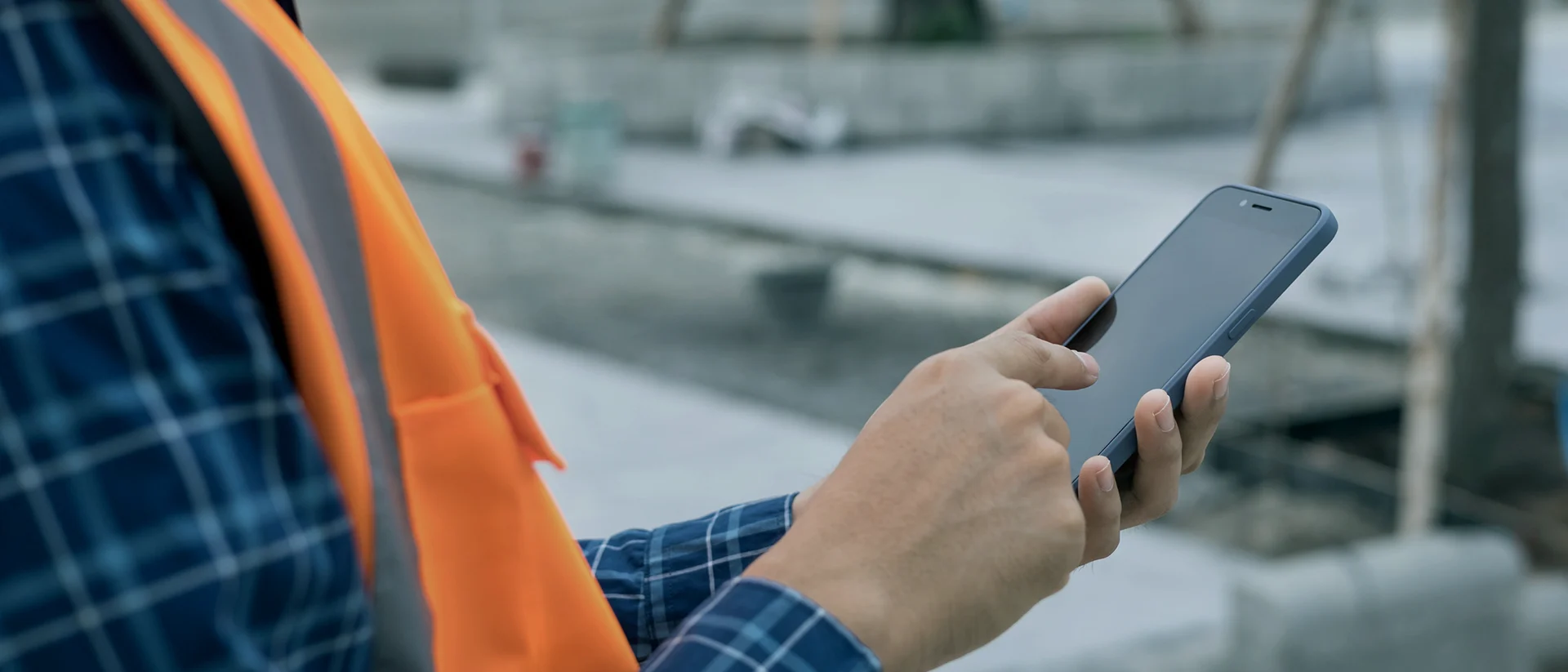 A person wearing an orange safety vest and plaid shirt uses a smartphone outdoors at a construction site, with only their hands and part of their torso visible.