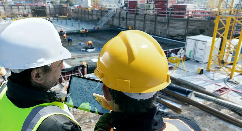 Two construction workers in hard hats look at a tablet displaying plans at a busy construction site, with several other workers and equipment visible in the background.