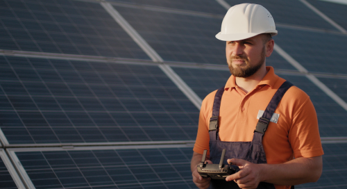 A man in an orange shirt and overalls wearing a white hard hat stands in front of solar panels, holding a remote control device.