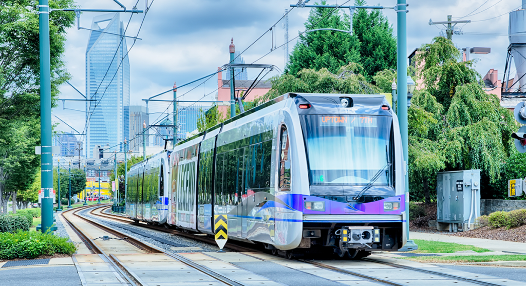 A modern light rail train travels on tracks through a city with tall buildings and trees in the background.