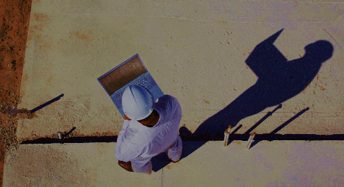 A person in a white hard hat and construction attire stands on a site, holding an open laptop, casting a clear shadow on the ground.