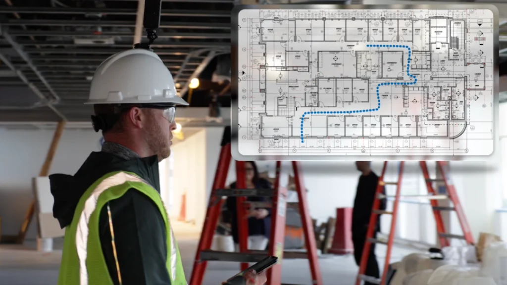 A construction worker in safety gear looks at a digital floor plan with a marked path, while standing in a partially finished building with ladders and exposed ceiling.