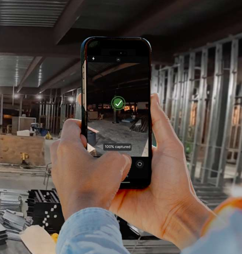 Person holding a phone, taking a photo of a construction site with metal framing; the phone screen shows a green checkmark and 100% captured, demonstrating the efficiency of construction reality capture software.