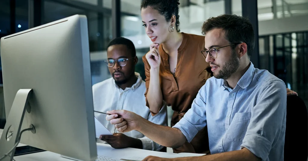 Three people work together at a desk, focused on a large computer monitor. One person points at the screen, while another holds papers and the third stands behind them, appearing engaged and collaborative.