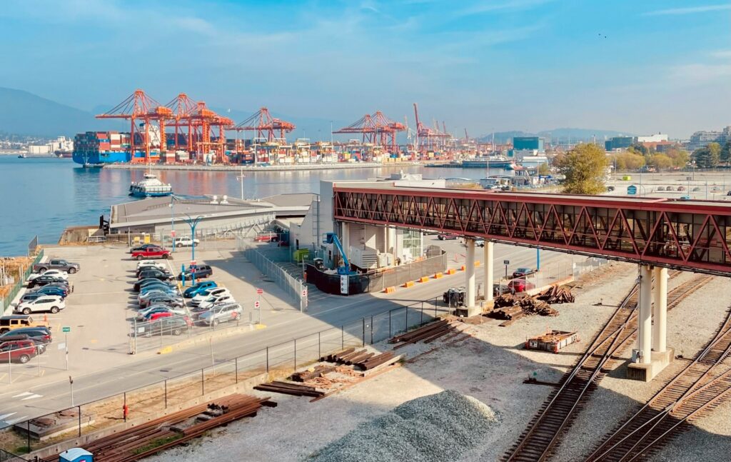 A busy shipping port with orange cranes, a parking lot, rail tracks, and a pedestrian bridge under a clear blue sky. Ships and containers are visible in the background.