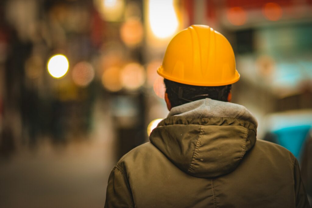 A person wearing a yellow hard hat and a green jacket stands with their back to the camera in a blurred urban setting with bright lights in the background.