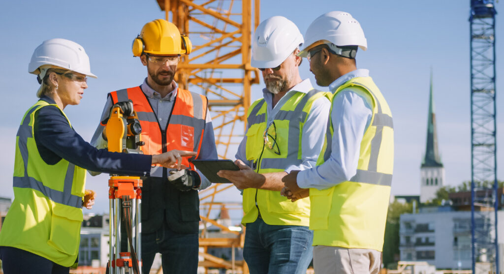 Four construction workers in safety gear stand on a site with cranes, reviewing information on a tablet. One person gestures to the screen while others observe and discuss. A church spire is visible in the background.