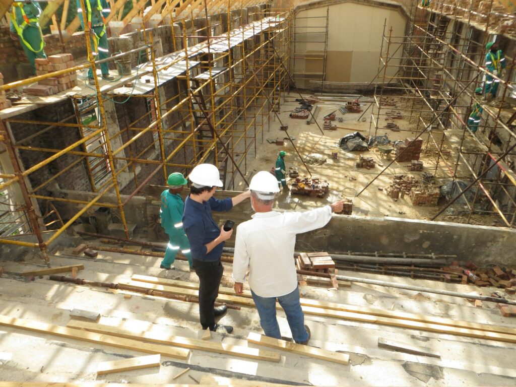 Two people in hard hats stand on a construction site, discussing plans. Scaffolding, wooden planks, and workers are visible around them as the building is being renovated or constructed.