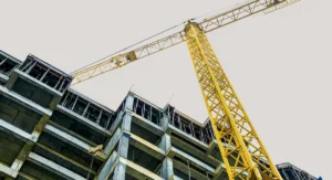 A yellow construction crane towers above a partially built, multistory concrete building against a pale sky, viewed from a low angle.