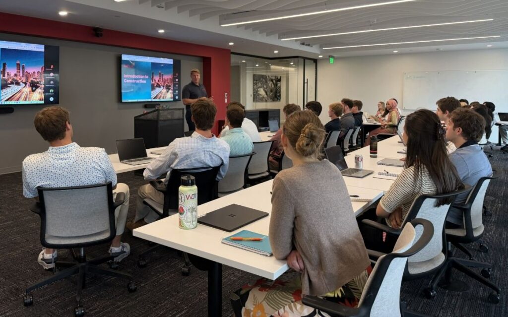 A group of people sits in a modern conference room listening to a presenter in front of slides titled Introduction to Construction. Tables have laptops and water bottles on them.