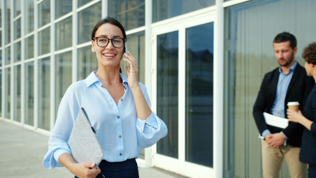 A smiling woman in a light blue button-down shirt and glasses talks on a smartphone outside an office building, while two colleagues talk in the blurred background.