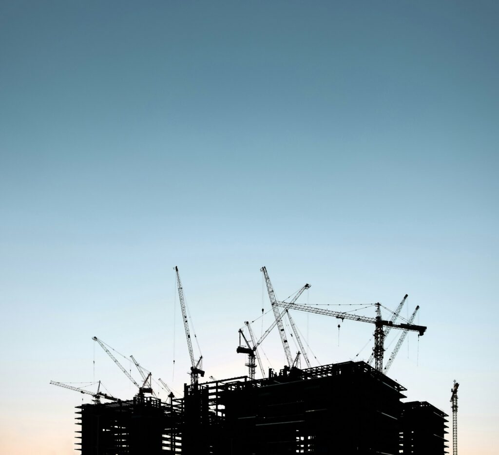 Silhouettes of multiple construction cranes towering over the framework of a building against a clear sky.