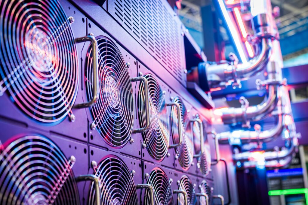 A row of cooling fans on a server rack illuminated with purple and blue light, with metal pipes extending into the background.