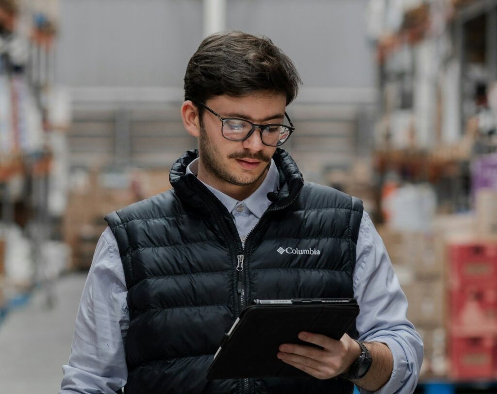 A man wearing glasses and a Columbia vest looking at a tablet in a warehouse.
