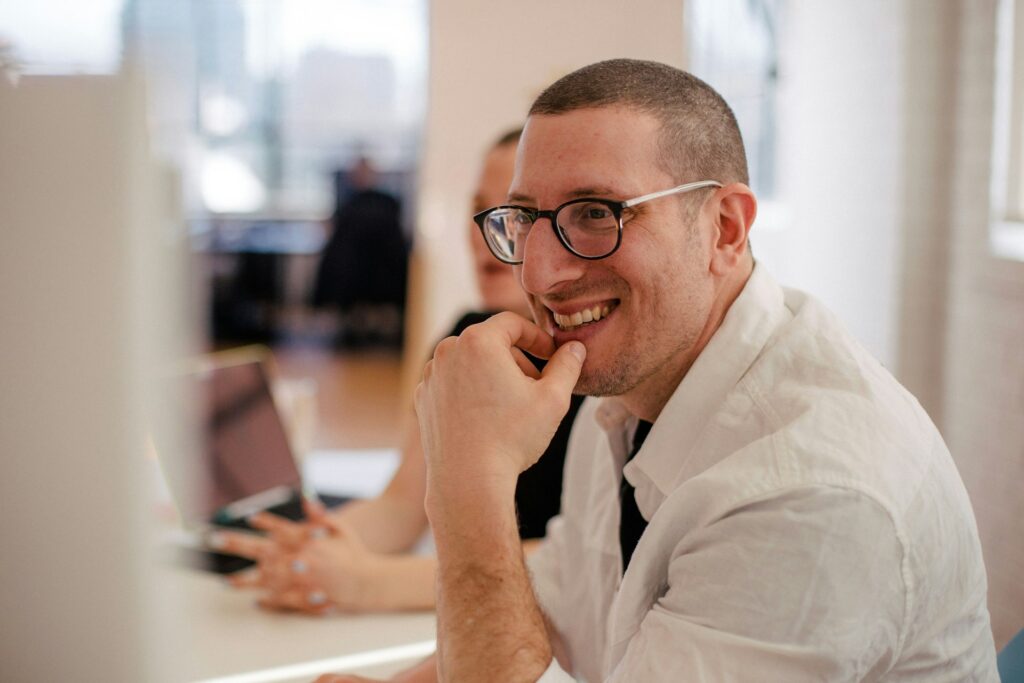 A man in a white shirt and glasses smiles while sitting at a desk with a laptop.
