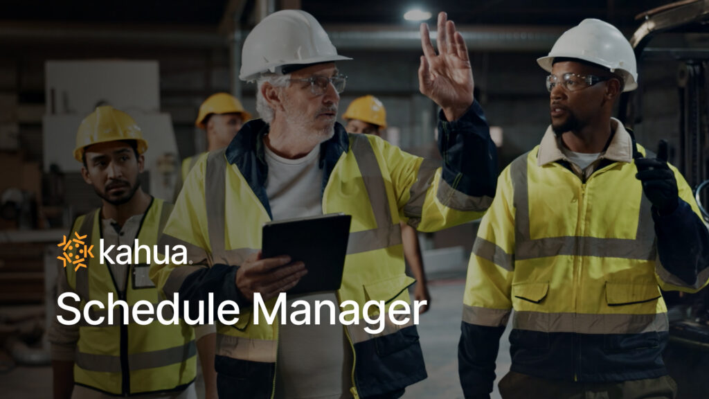 Construction workers in hard hats and high-visibility vests use construction scheduling software on a tablet in a workspace with the Kahua Schedule Manager logo overlaid.